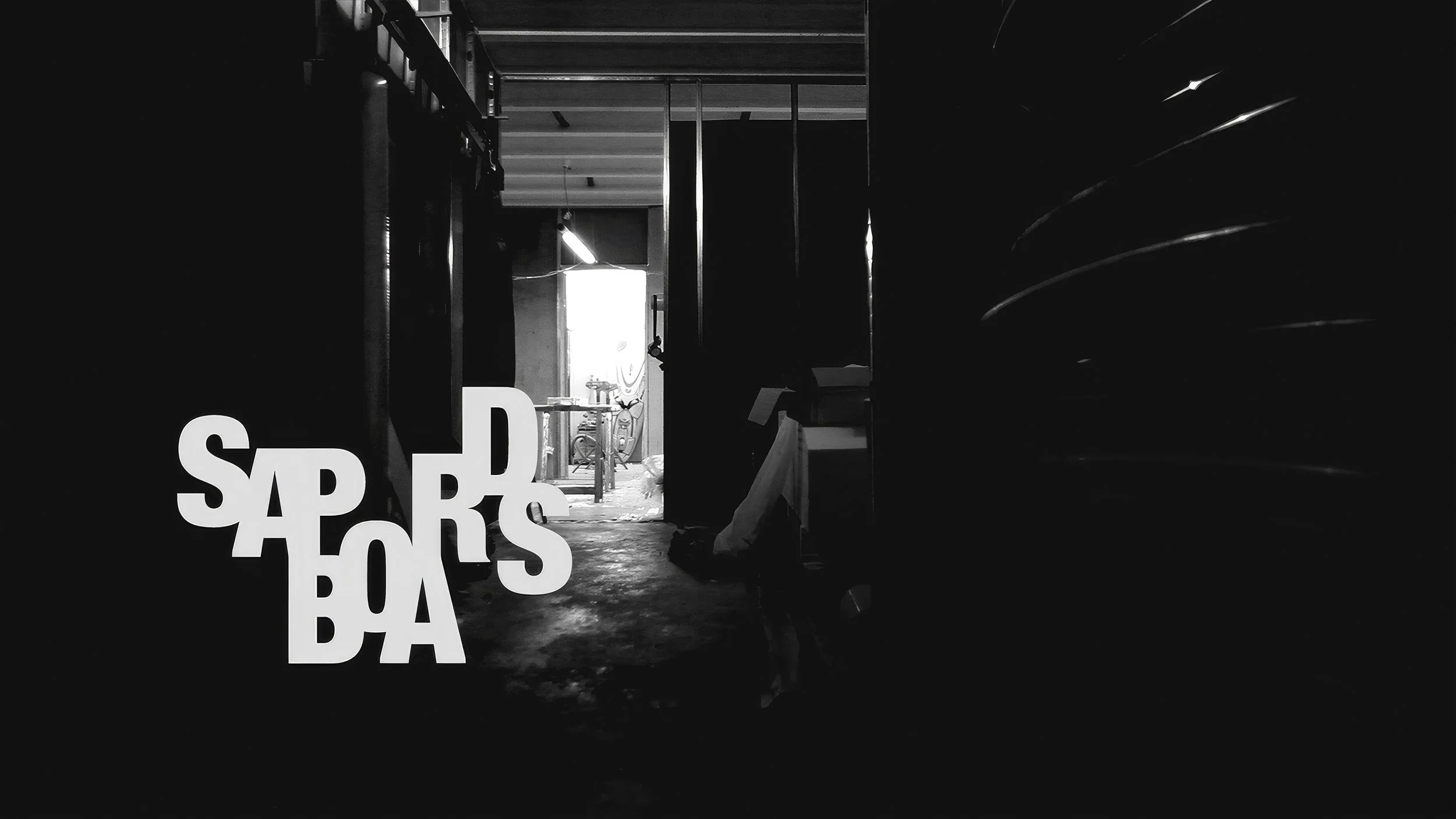 Black and white photograph of an industrial space with a door opening to a brightly lit room with bicycles and tools. The foreground features large, overlapping white letters spelling out "SPAR BOTA."