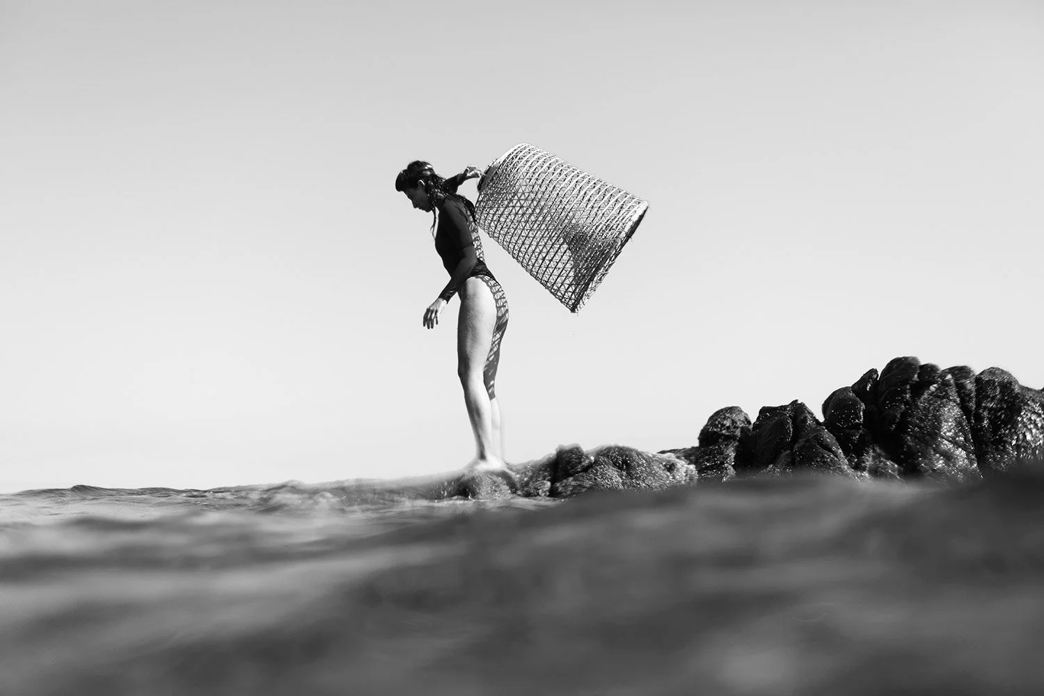 A woman standing on rocks at the edge of the water, carrying a laundry basket on her shoulder, with the horizon and sky in the background, in black and white.