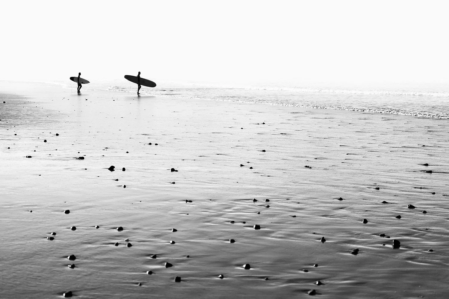 Two surfers walking along the shoreline carrying surfboards on a calm beach, black and white photo.