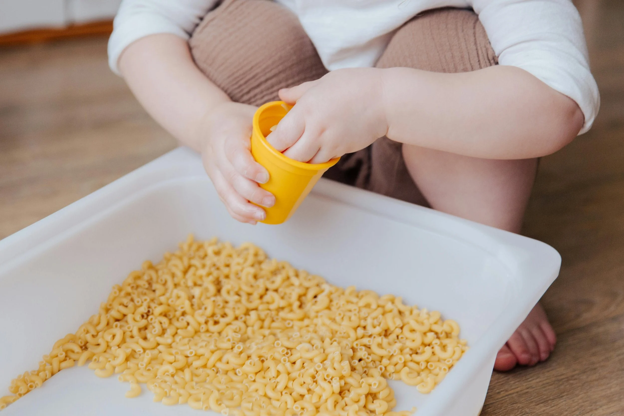 Child pouring dry elbow macaroni into a white tray using a yellow cup, sitting on a wooden floor.
