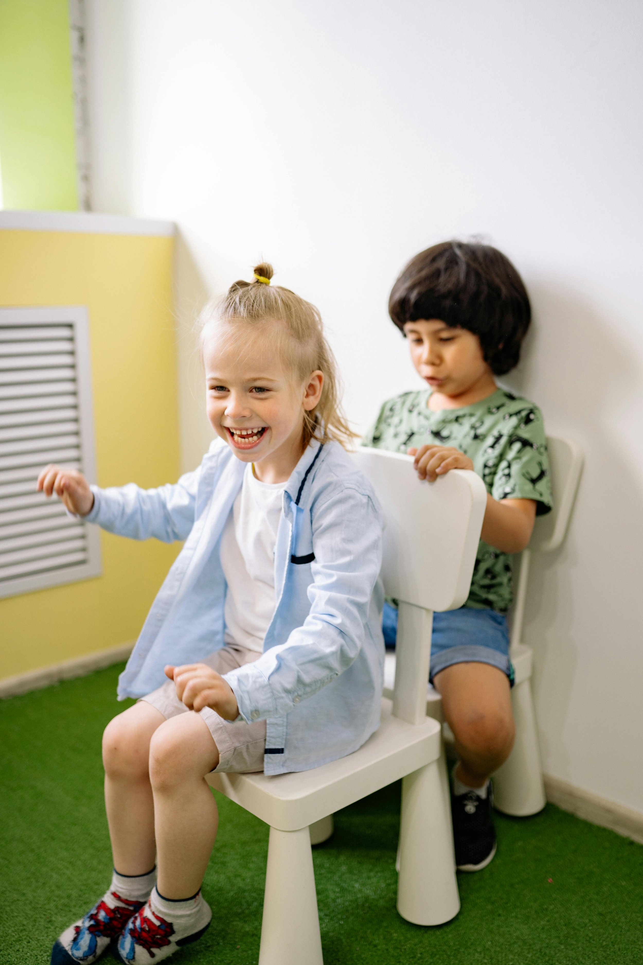 Two children, a girl and a boy, sitting on chairs against a white wall, with the girl laughing and the boy looking down at a tablet in his hands.