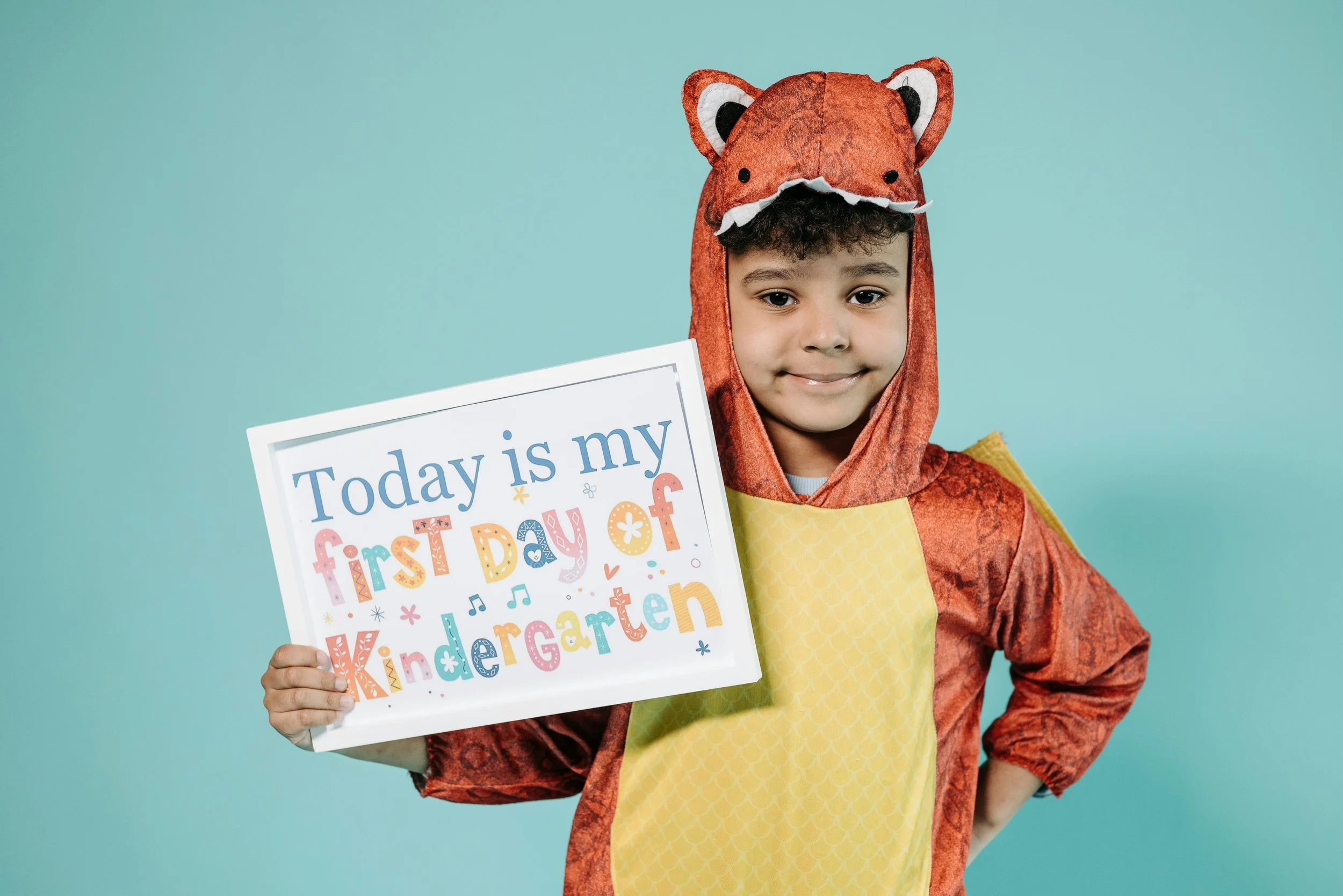 Young boy dressed in an animal costume holding a sign that reads 'Today is my first day of Kindergarten' against a light blue background.