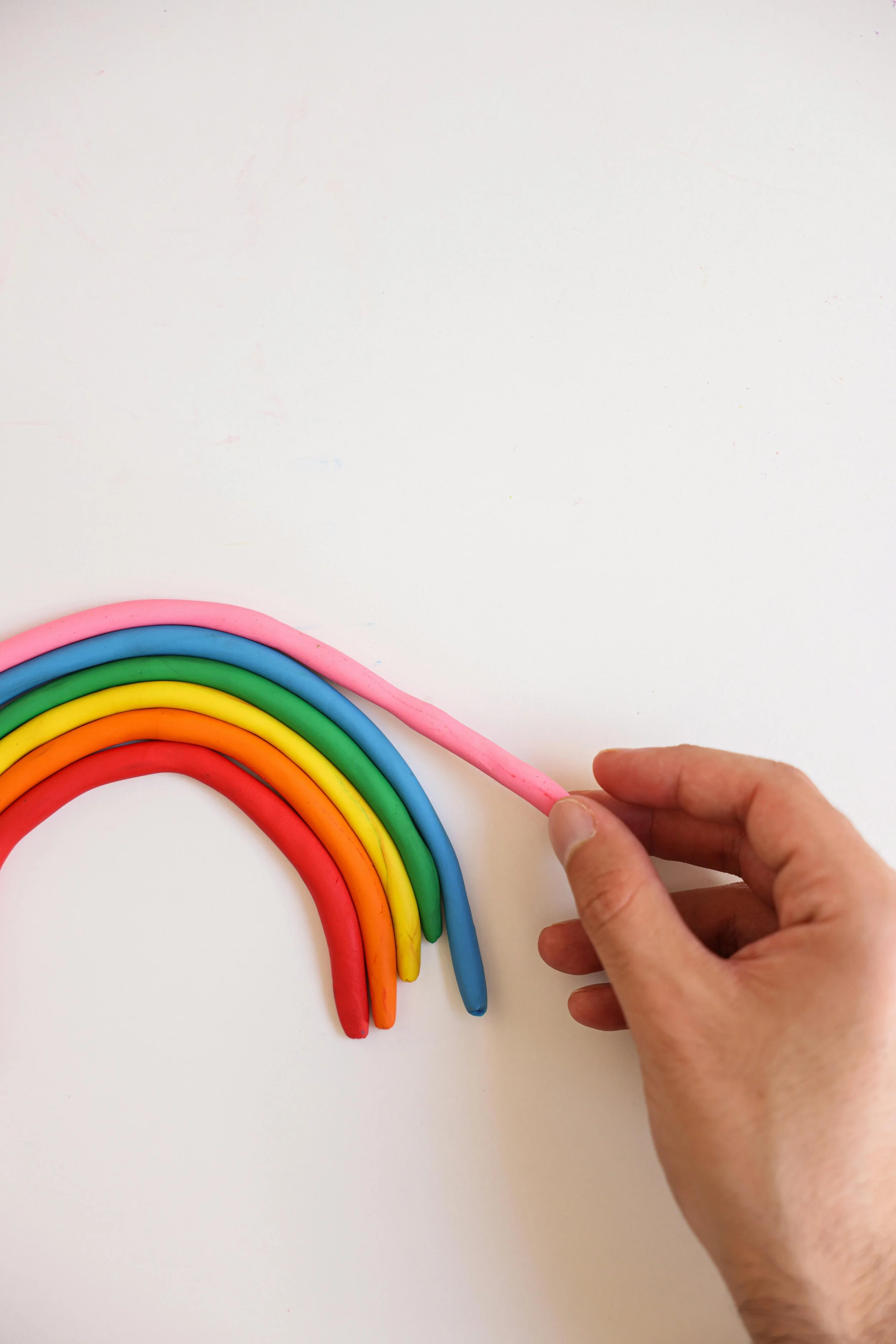 Hand holding pink pipe cleaner over a rainbow of colorful pipe cleaners on a white surface.