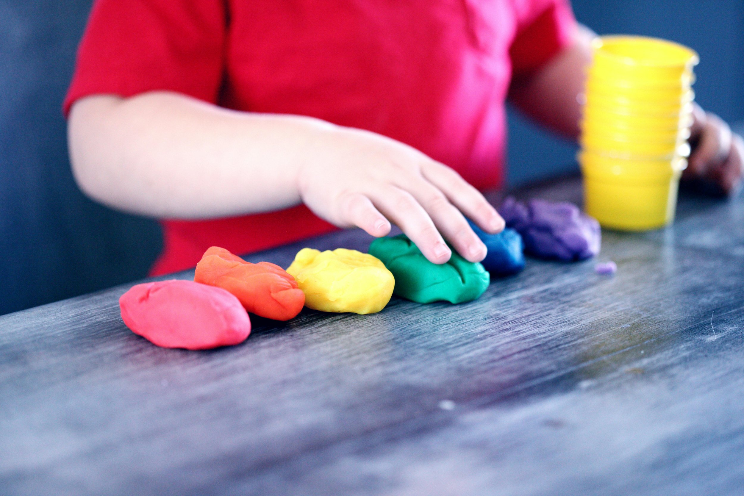 A child wearing a red shirt playing with colorful playdough on a dark wooden table, with a stack of yellow containers in the background.
