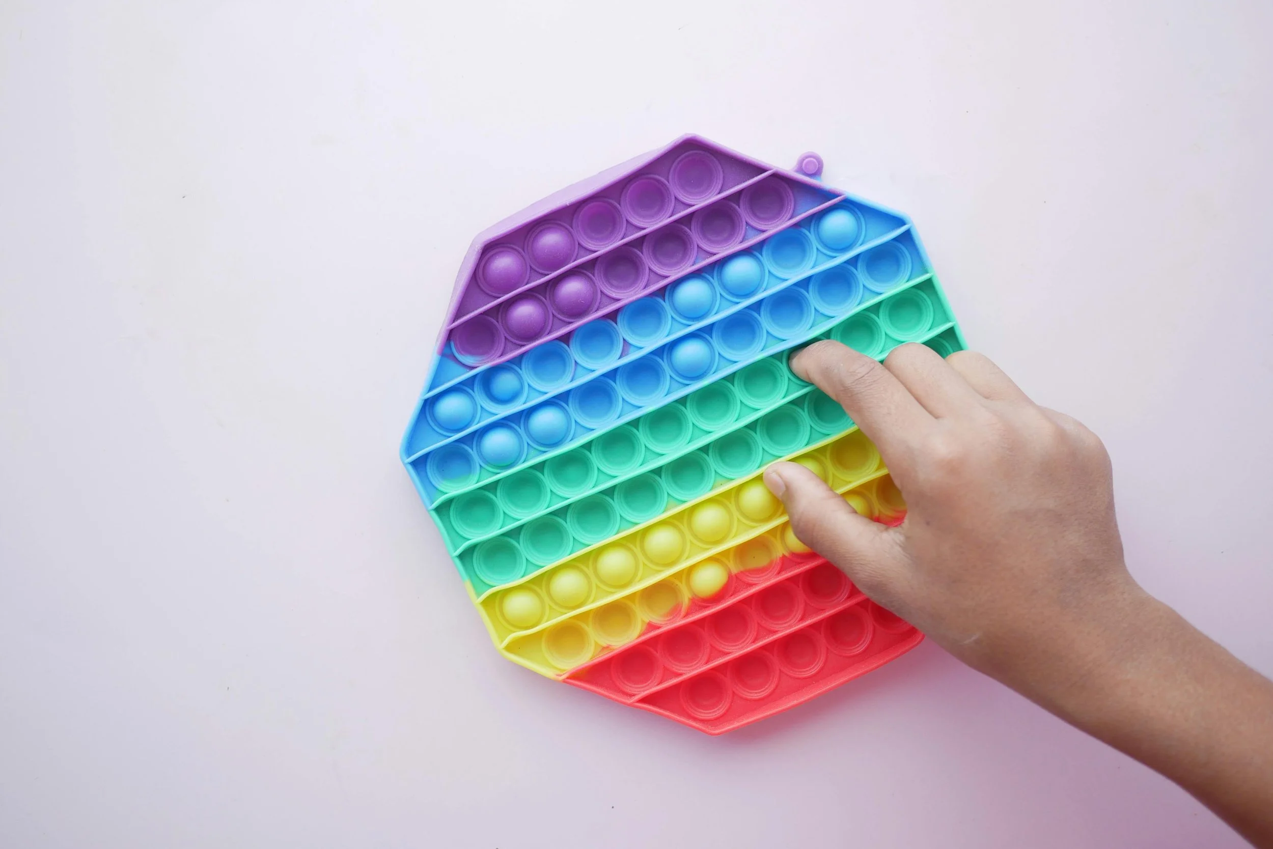 A hand pressing on a colorful, hexagonal-shaped pop it fidget toy with rainbow colors on a white surface.