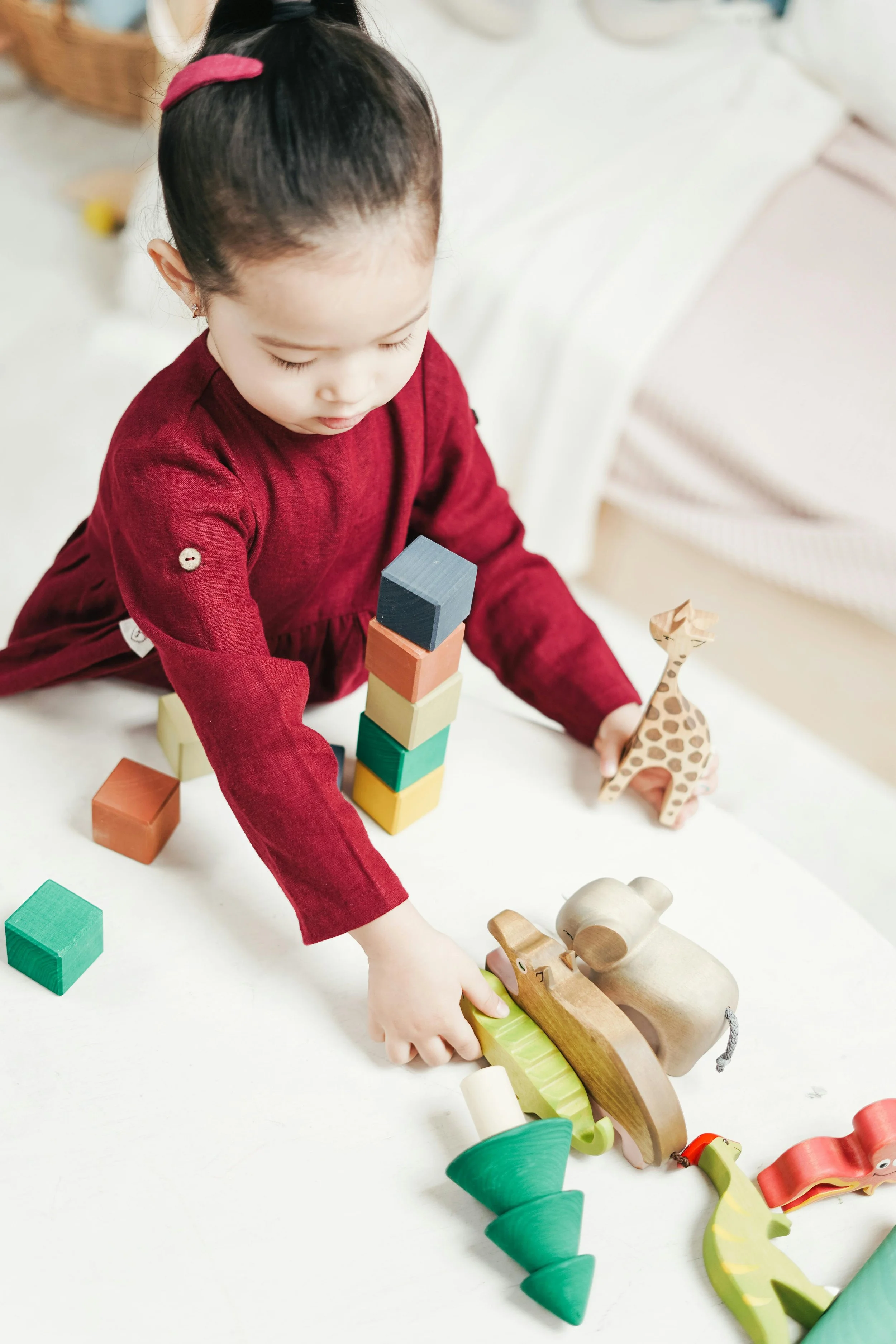 A young girl in a red long-sleeve shirt playing on a white table with wooden animal toys and building blocks.