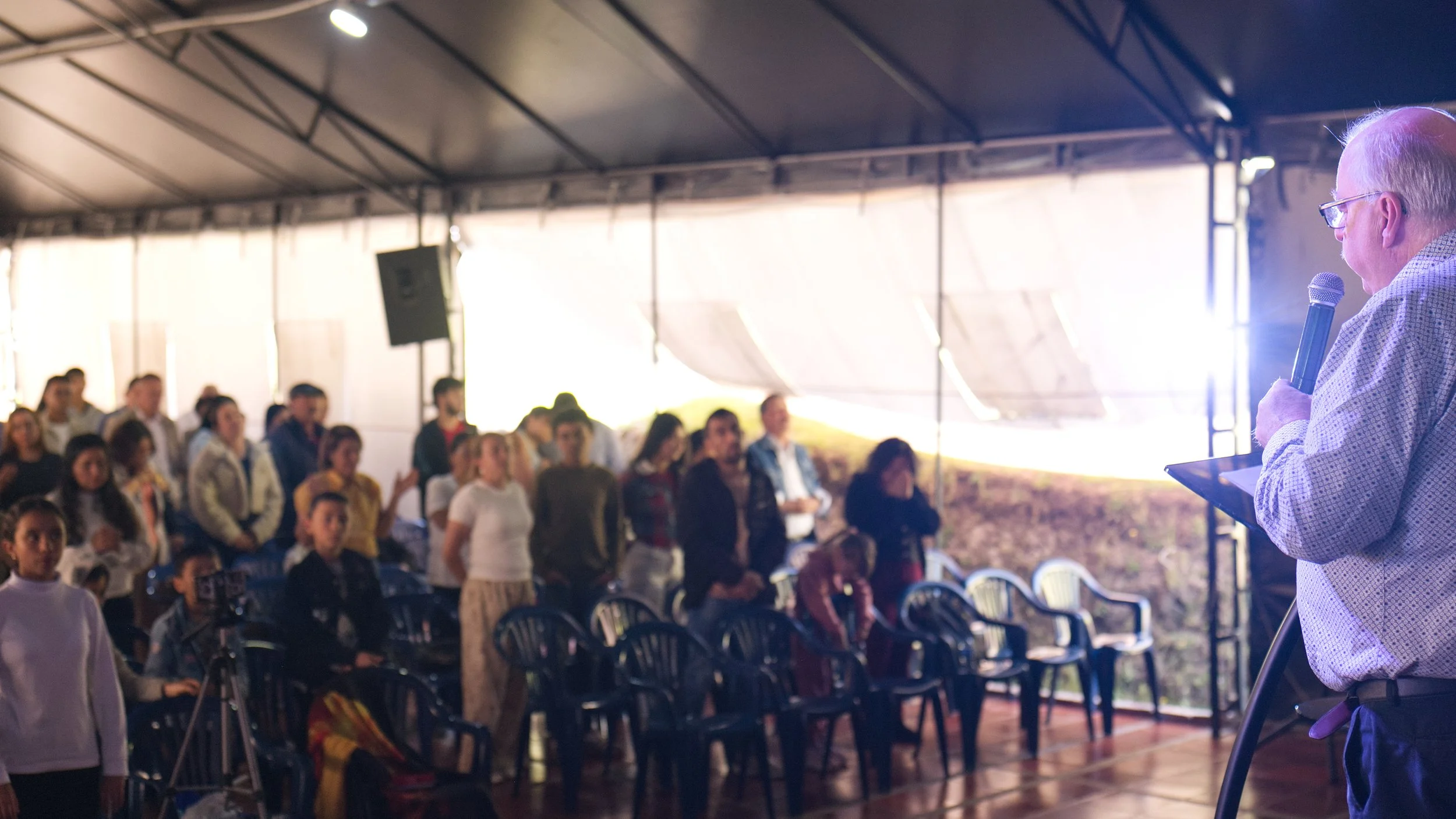 A man with glasses and a light-colored shirt holding a microphone and speaking to an audience of diverse people standing and sitting in a large tent.