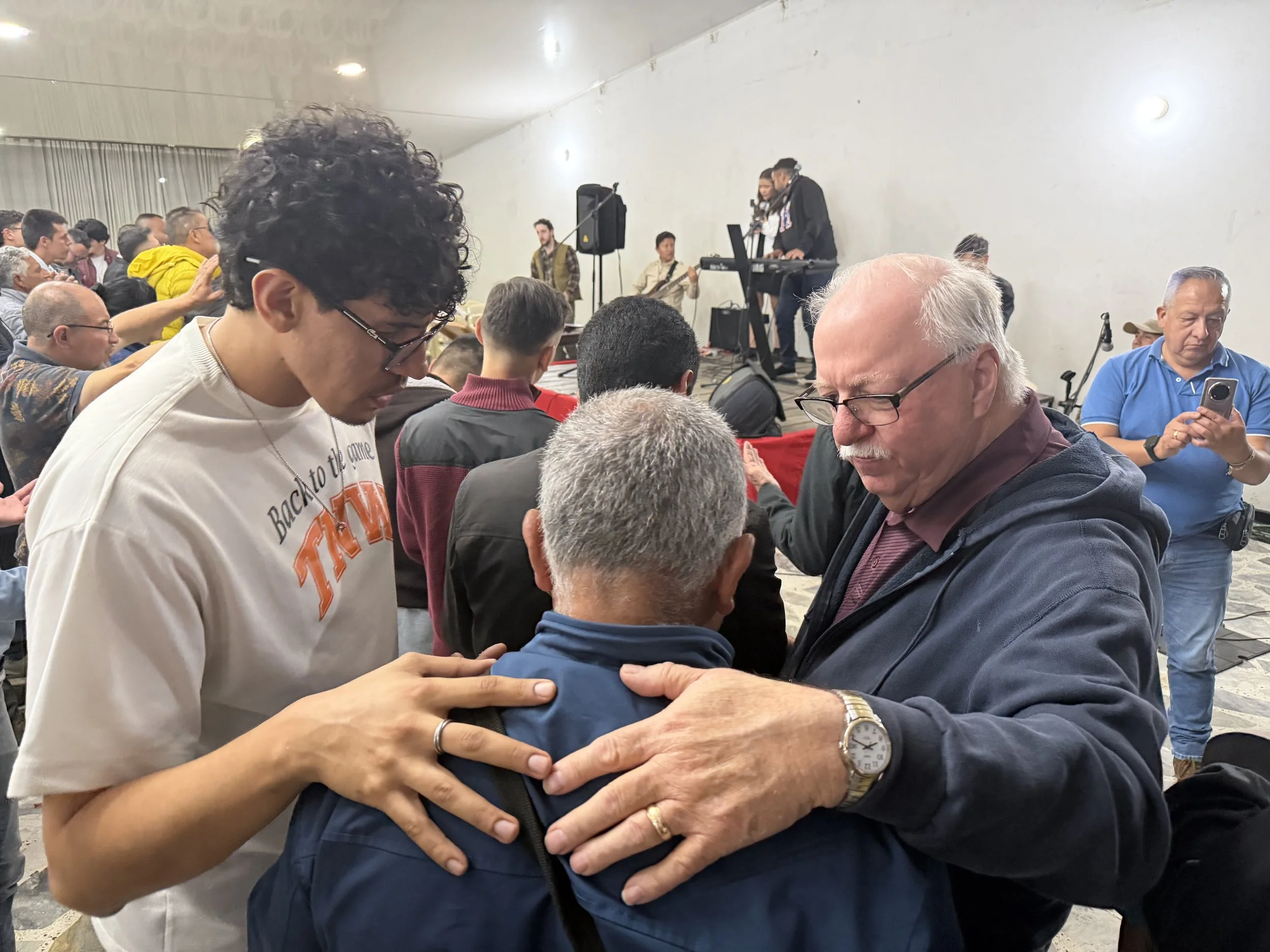 A diverse group of people gathered indoors, with three individuals in the foreground placing their hands on a person's shoulder, during what appears to be a prayer or blessing. In the background, a small band performs on a stage, and other attendees are either participating, observing, or taking photos.