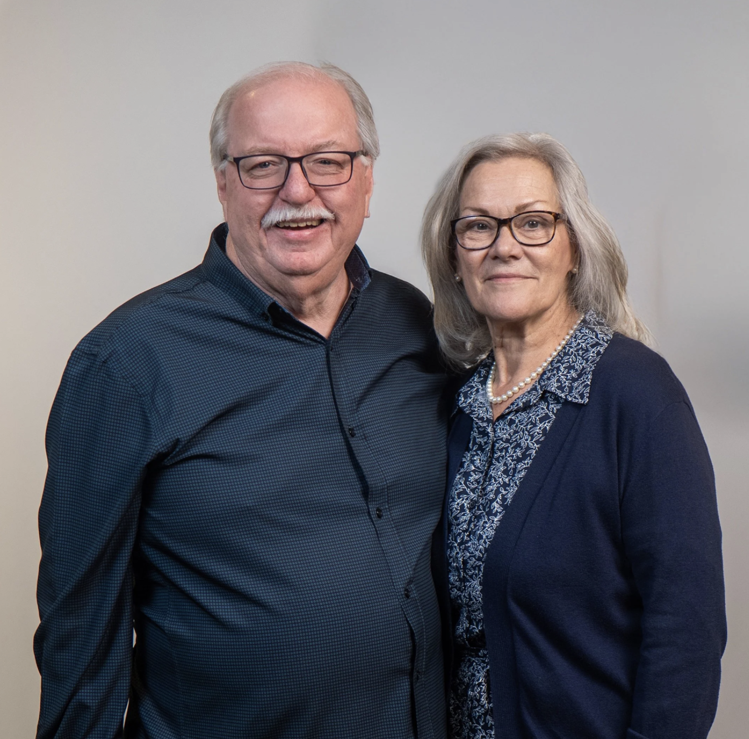 A smiling elderly man with glasses and a mustache, standing next to an elderly woman with glasses and a pearl necklace, both wearing dark blue clothing against a light grey background.