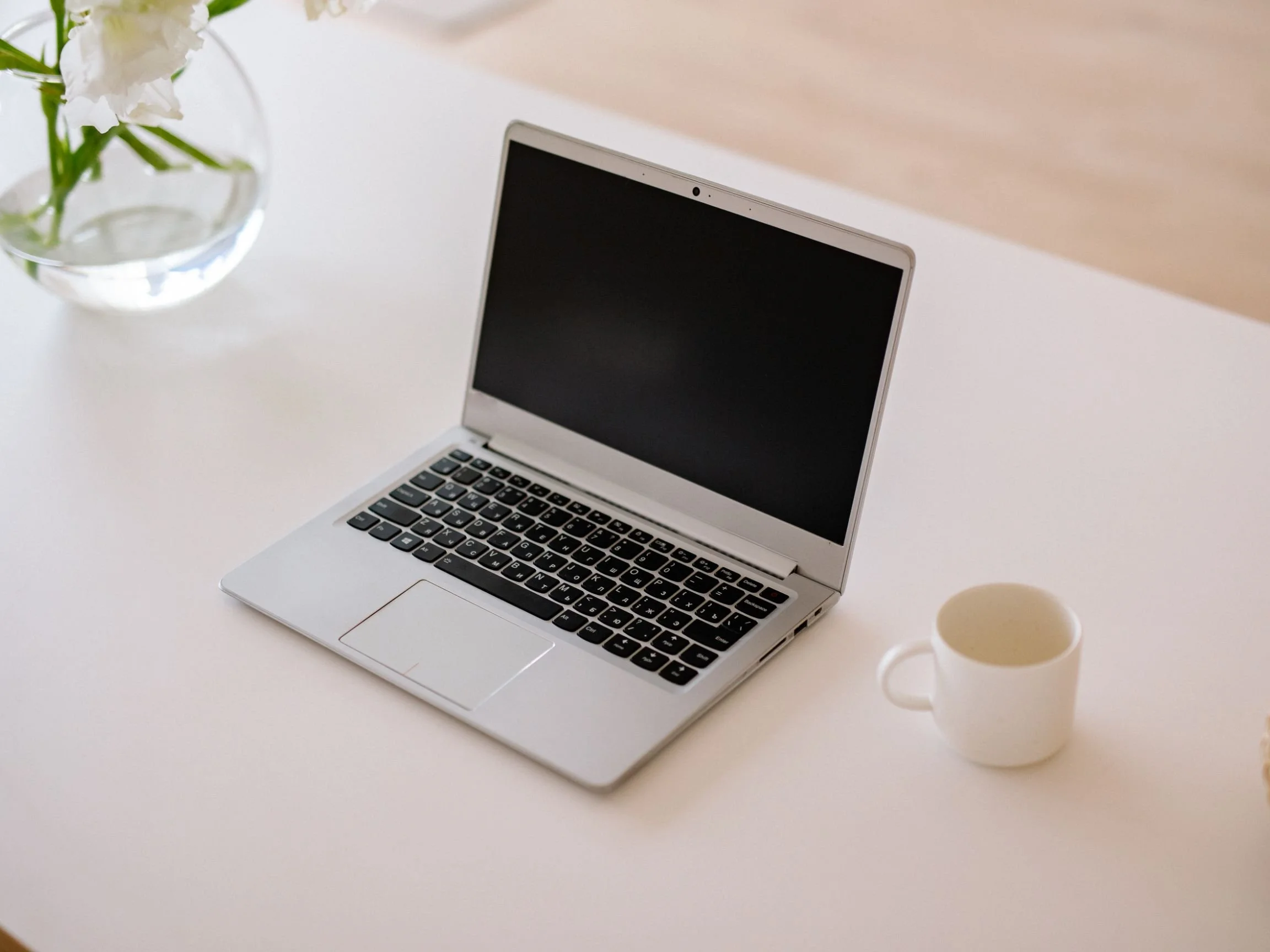 Open silver laptop with black keyboard and blank screen on a white desk next to a white coffee mug and a glass vase with white flowers.