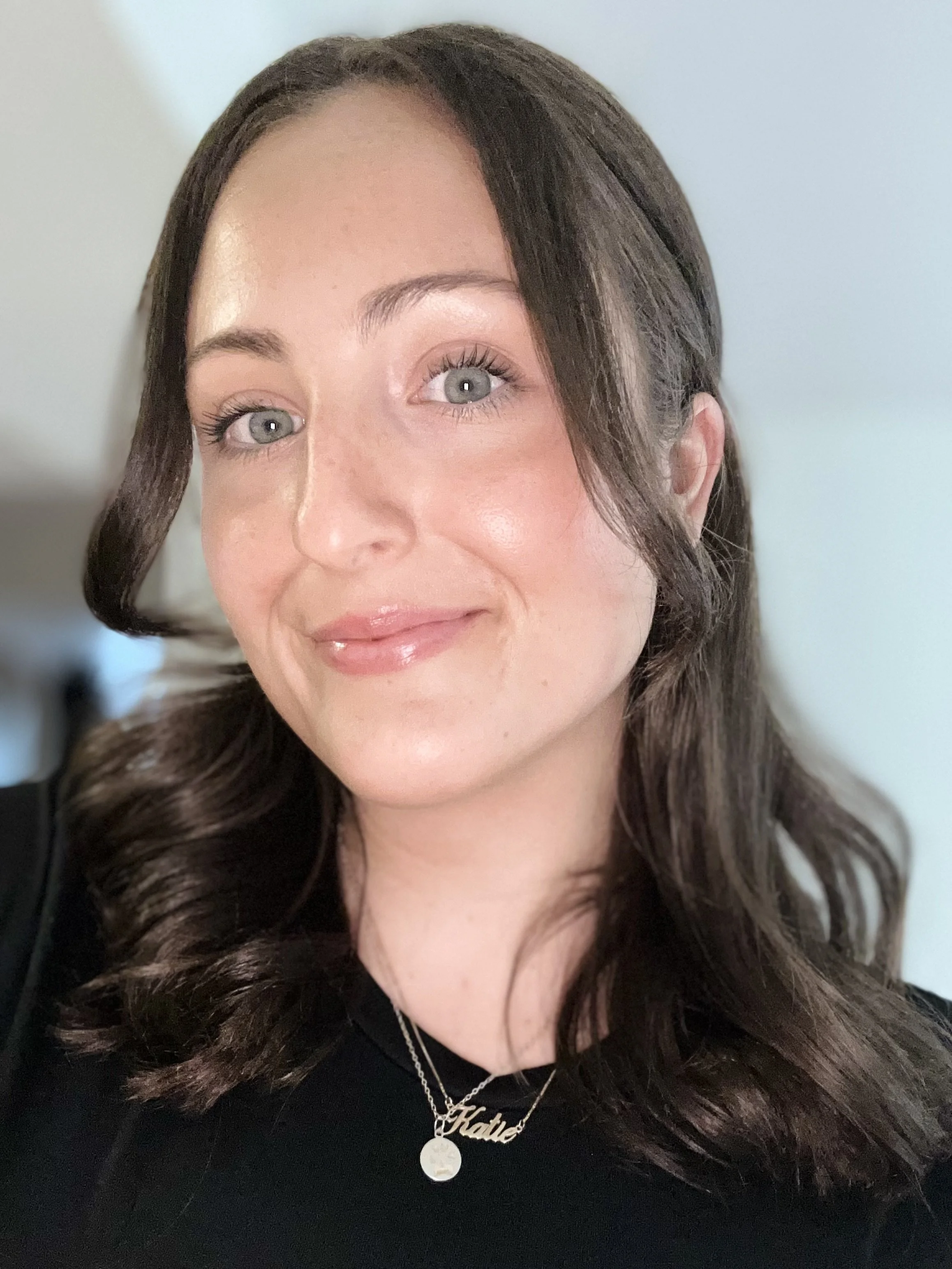 Close-up of a woman with shoulder-length wavy brown hair, blue eyes, and light makeup smiling at the camera, wearing a black top and layered necklaces.