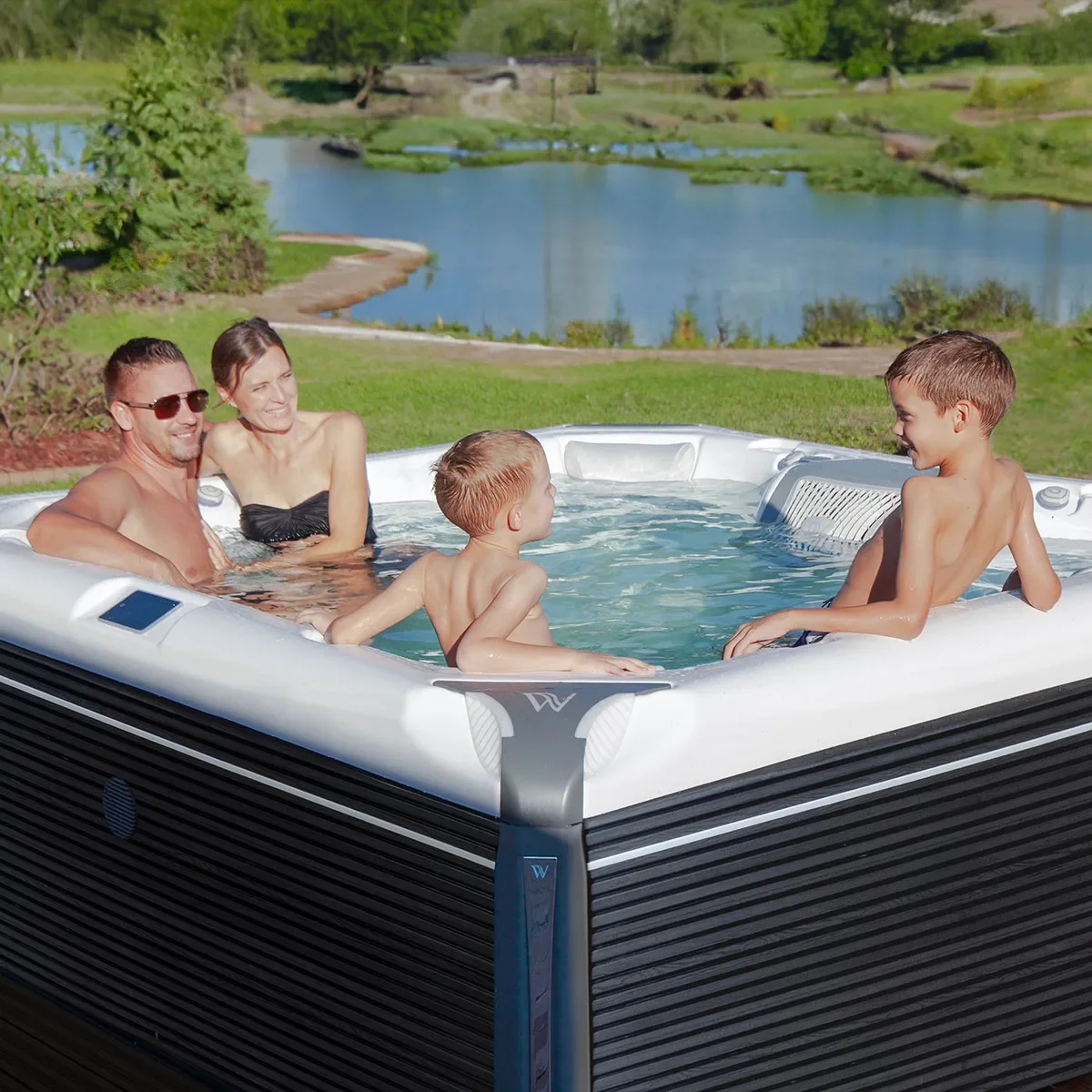 A family of four enjoying a hot tub outdoors near a pond on a sunny day.