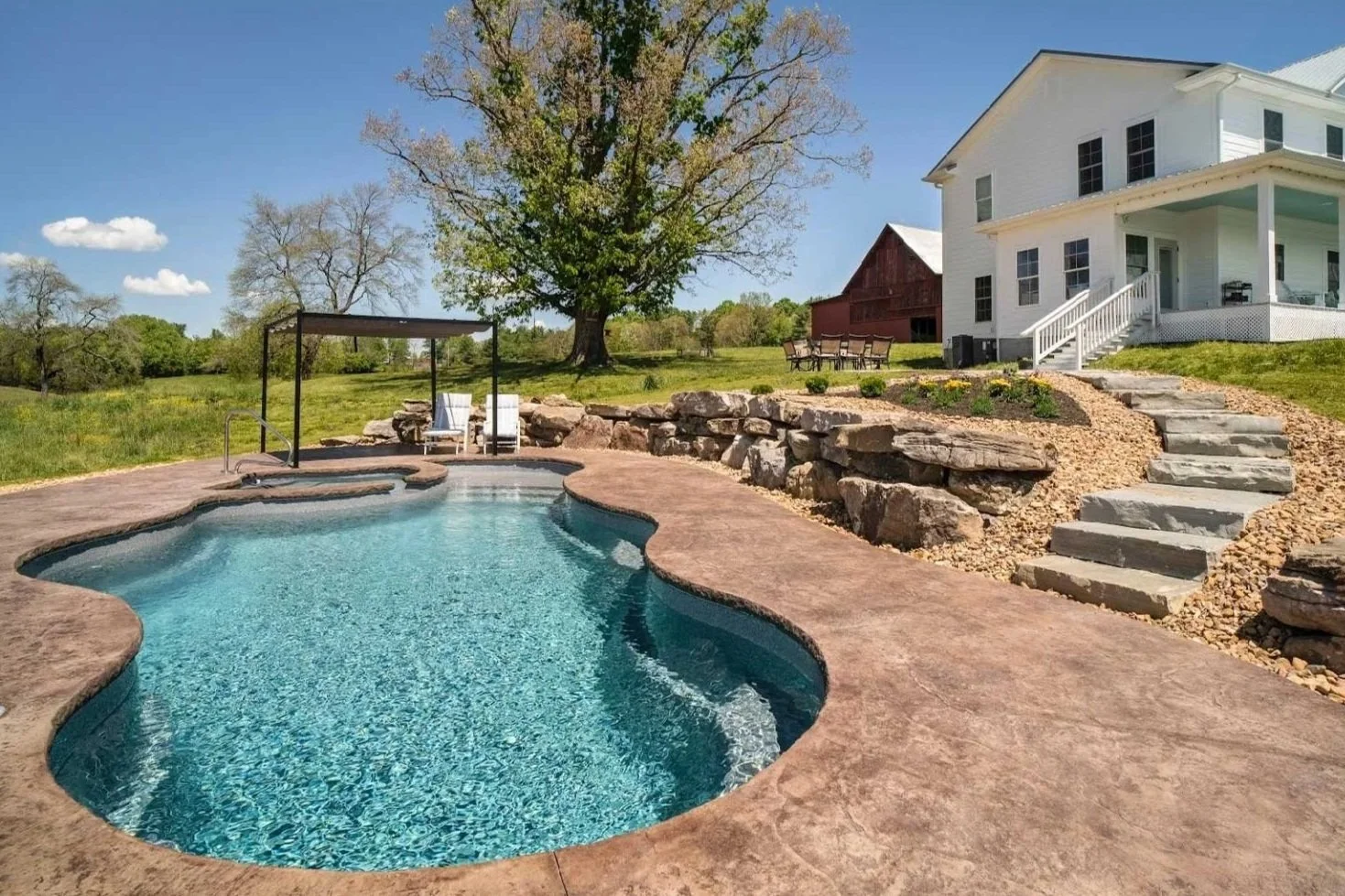 A backyard with a swimming pool, stone steps, a white house, outdoor seating, a large tree, and a clear blue sky.