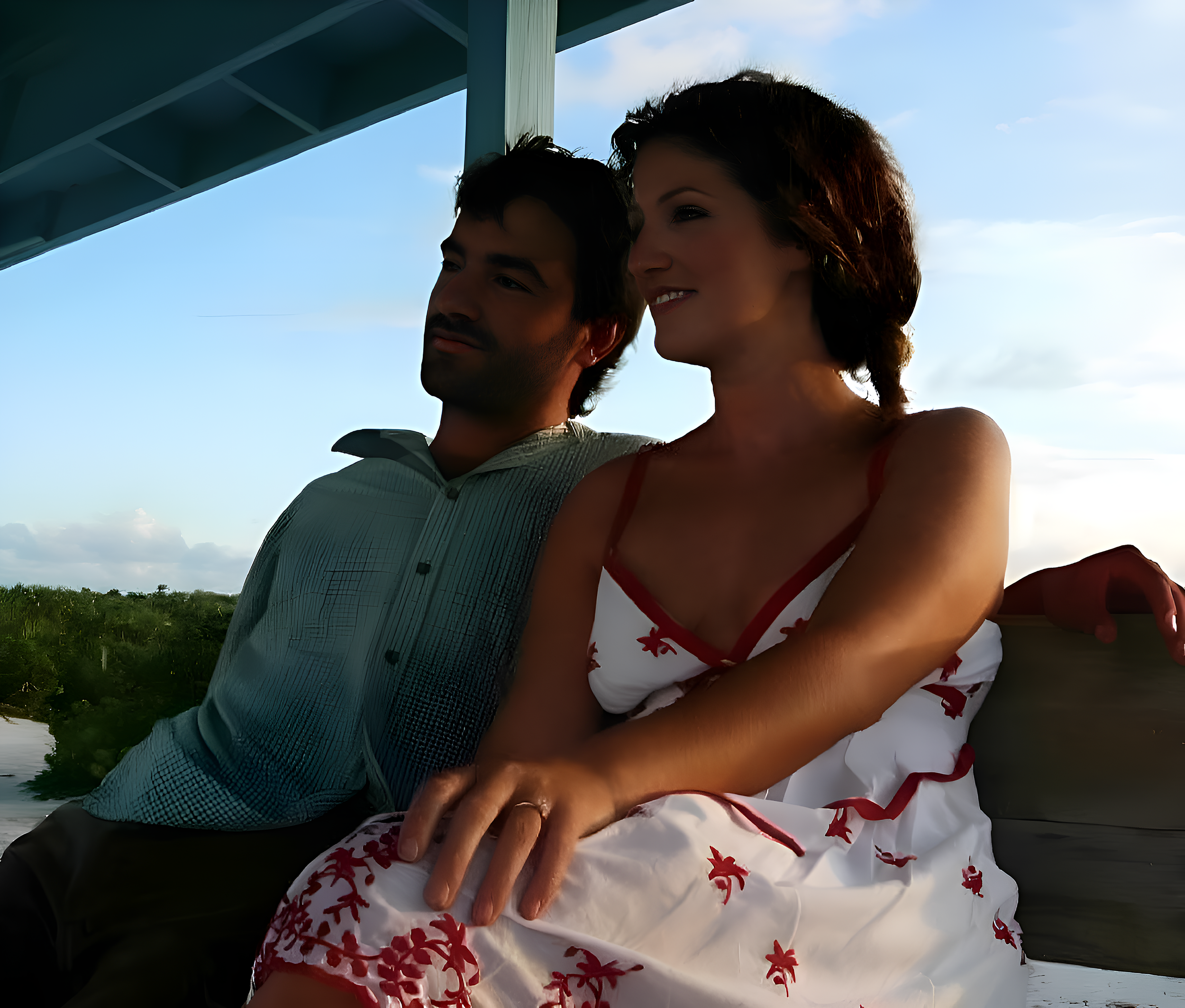 A man and woman sitting close together outdoors under a shelter, with a cloudy sky and greenery in the background, appearing relaxed and happy.