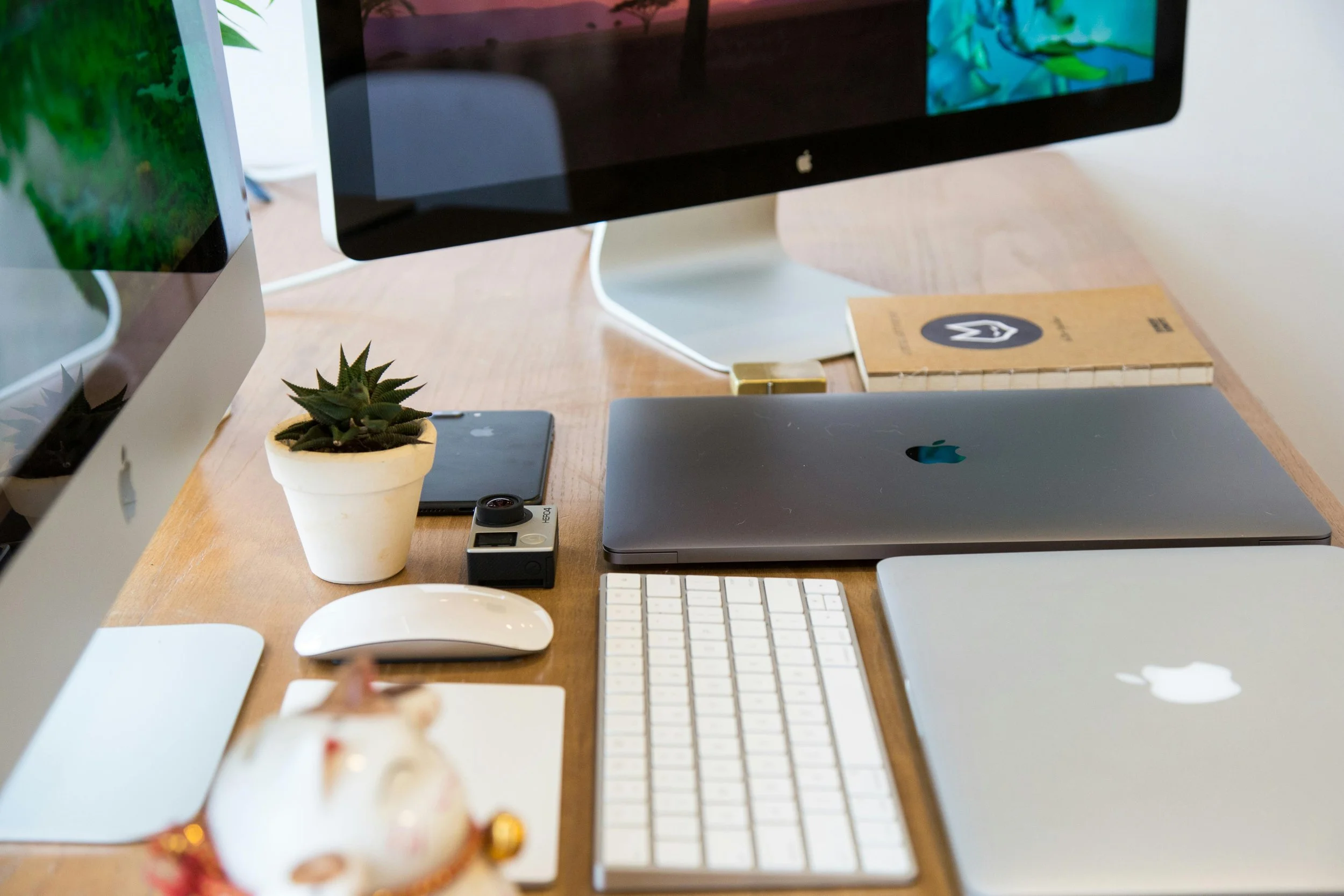 Desk with two monitors, a laptop, a keyboard, a mouse, a potted plant, a smartphone, an action camera, a notebook, and a small decorative teapot.