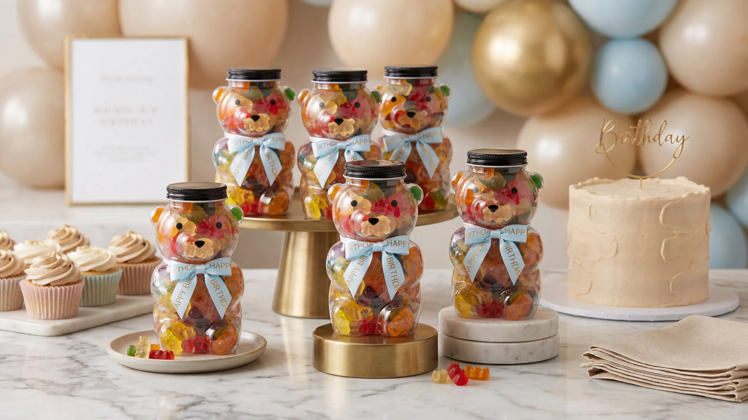 Candy jars with bear face designs, cupcakes with swirled frosting, and a birthday cake with a "Birthday" topper on a marble table.