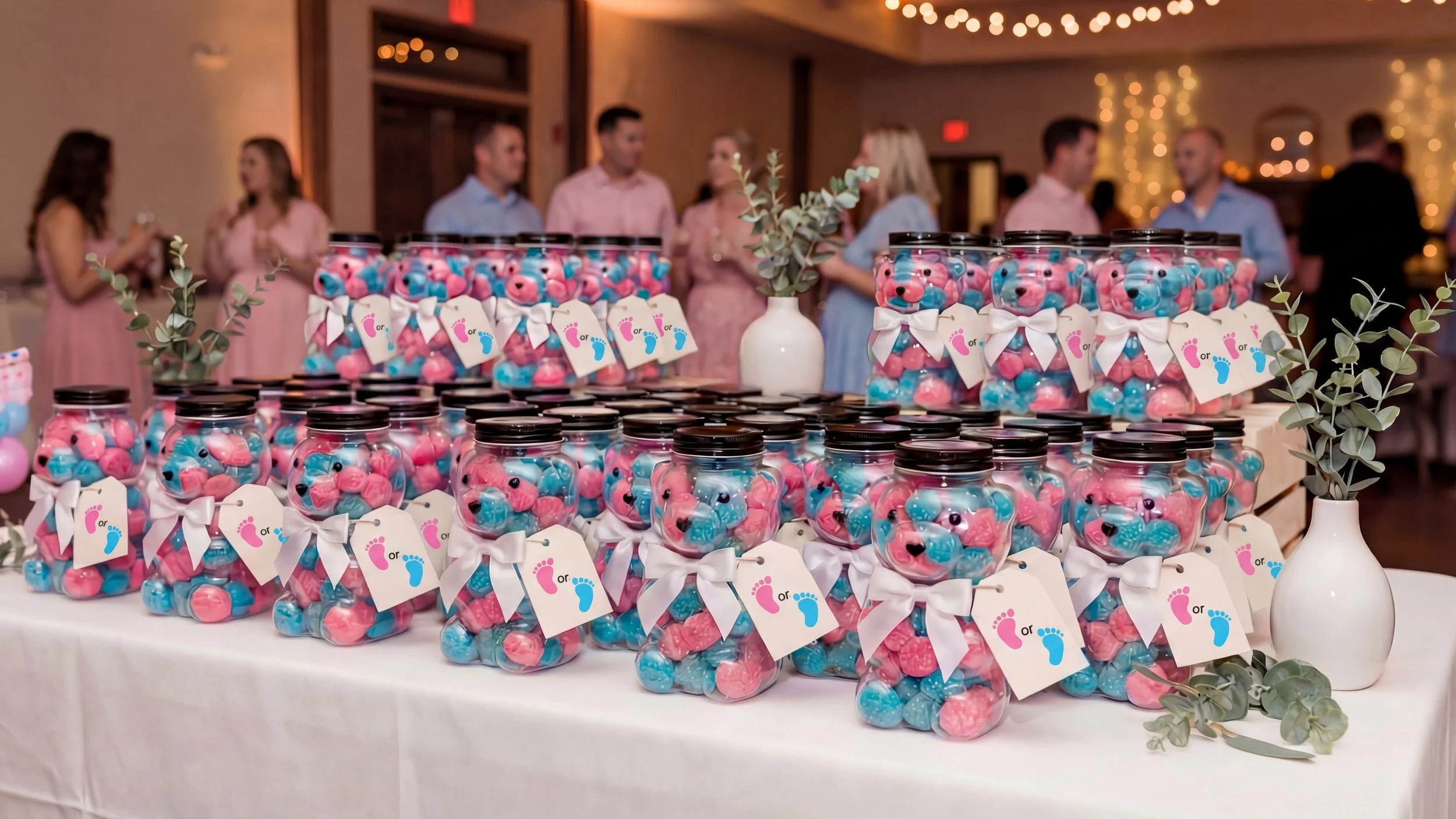 Table decorated for a baby shower with jars filled with pink and blue candies, each tied with a white ribbon and tags with pink and blue footprints, and small vases with greenery in the background, with people socializing in the background.