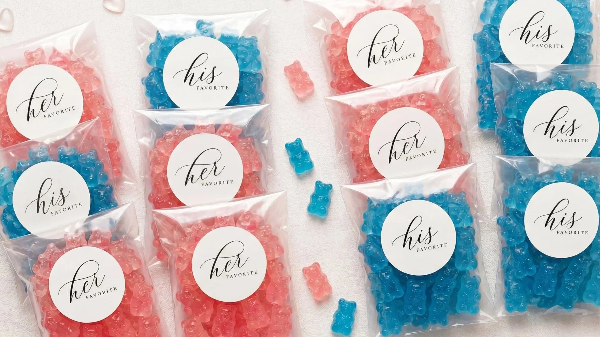 Small gummy bear candies in pink and blue, packaged in clear bags with white circular labels that read 'her favorite' and 'his favorite' in elegant black script.