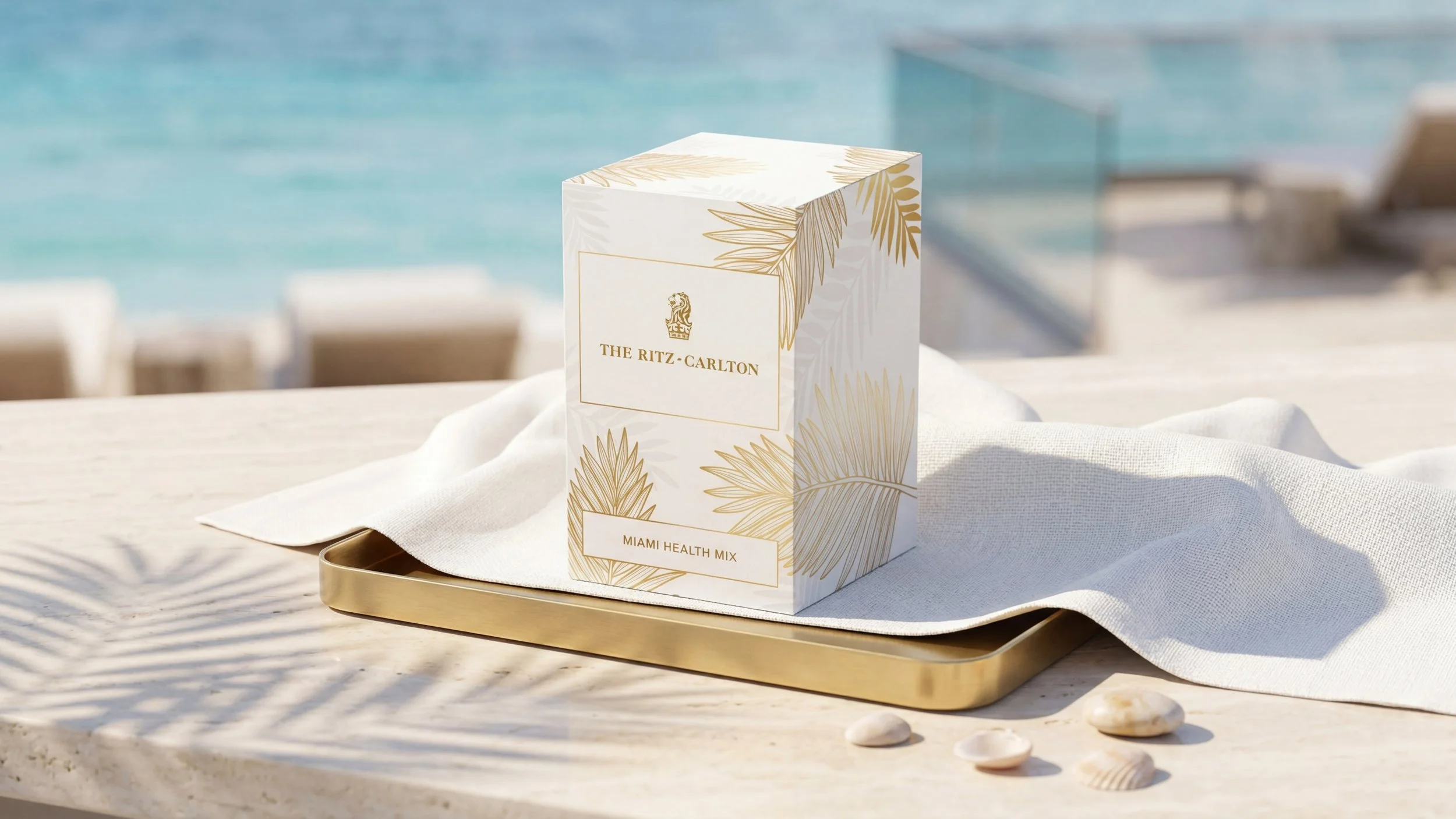 A box of Miami Health Mix by The Ritz-Carlton sits on a gold tray, draped with a white cloth, with seashells on a table, by a pool.