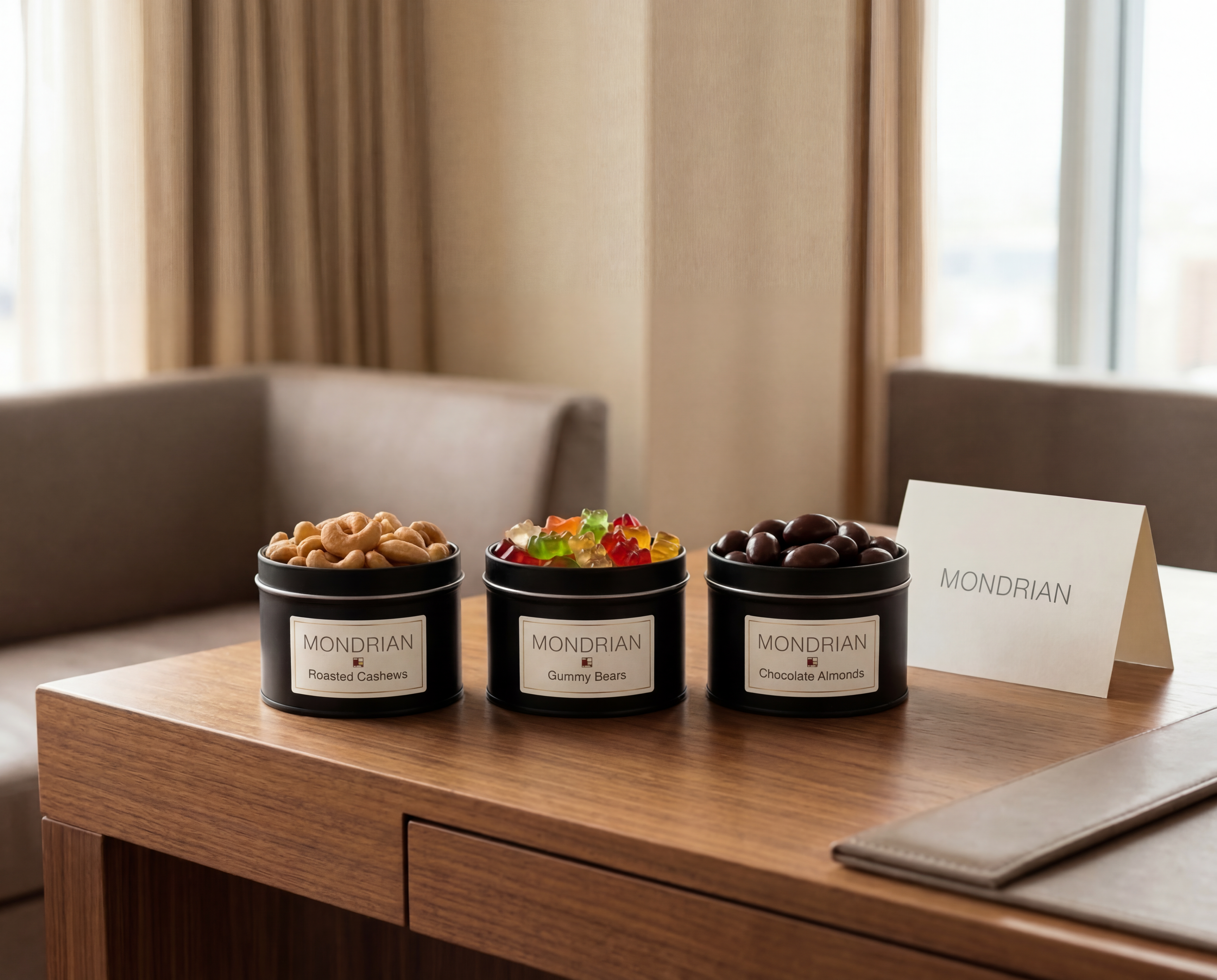 Three black containers with labels holding assorted candies and nuts on a wooden table in a hotel room, with a white sign and beige curtains in the background.
