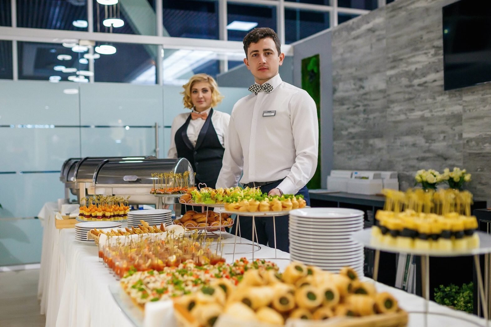 Coadiuvanti in uniforme con papillon, tra un tavolo di antipasti e finger food, in un ambiente moderno e luminoso.