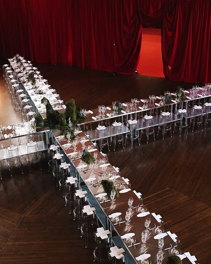 Una sala da pranzo elegantemente allestita con lunghi tavoli con tovaglie bianche, piatti, bicchieri e posate, decorati con fiori e piante verdi, pronta per un evento o una festa, con un palco sullo sfondo con tende rosse.