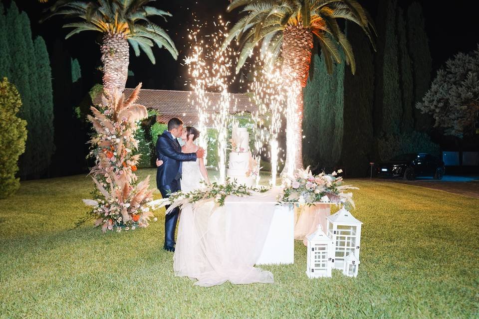 Un matrimonio che danza all'aperto di notte, decorazioni floreali e fuochi d'artificio, con palme e alberi sullo sfondo