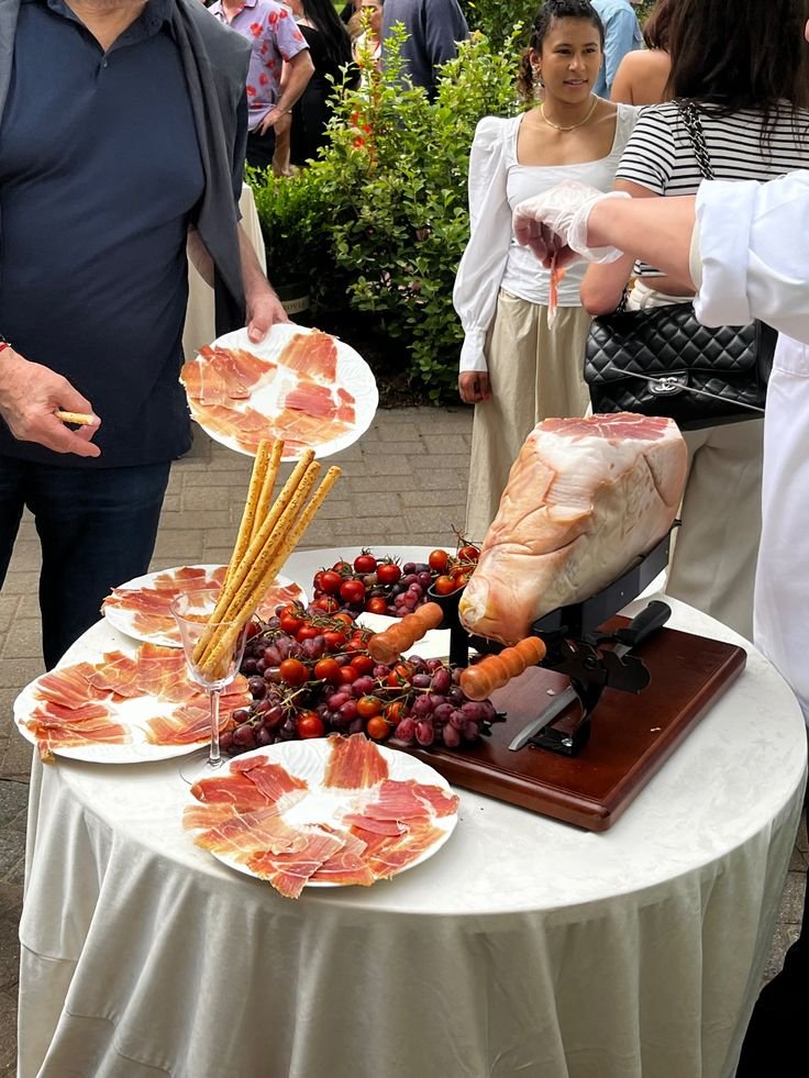 Tavolo con prosciutto impanato, uva, ciliegie e un prosciutto in preparazione, con persone che stanno degustando e acquistando i prodotti.
