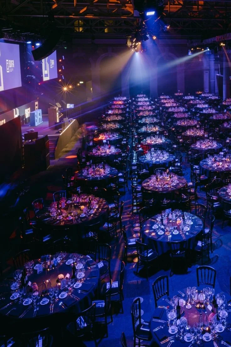 Sala elegante allestita con tavoli rotondi apparecchiati per un evento, illuminata da luci colorate e luci da discoteca, con un palco e uno schermo sullo sfondo.