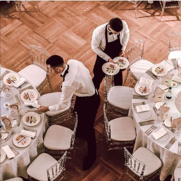 Camerieri in abiti formali che preparano tavoli con piatti e posate elegantemente apparecchiati.