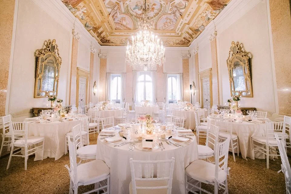 Sala da pranzo elegante con tavoli rotondi e sedie bianche, decorazioni floreali, grandi specchi dorati, pareti e soffitto decorati con affreschi e un grande lampadario di cristallo al centro.