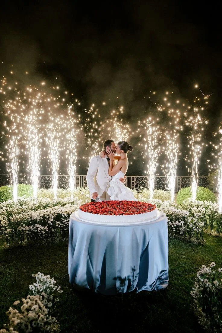 Una coppia di sposi si bacia davanti a una torta nuziale decorata con frutta, con fuochi d'artificio e luci scintillanti sullo sfondo, durante una celebrazione all'aperto di sera.