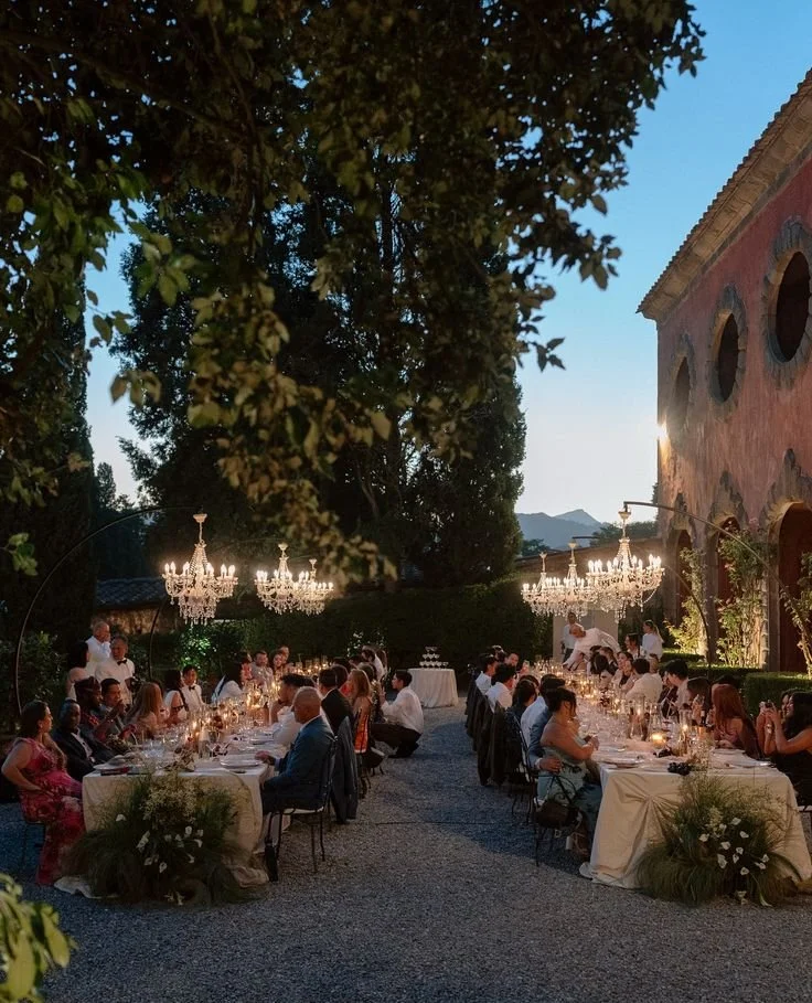 Cena all'aperto con tavoli apparecchiati, ospiti seduti, ortamte illuminato da chandelier, al tramonto in un cortile con alberi e un edificio storico.