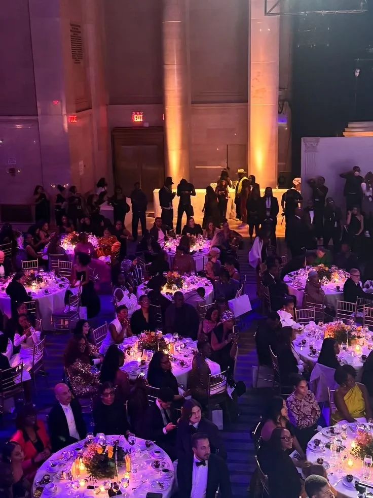 Una sala elegante con tavoli decorati per un evento formale, con molte persone sedute e altre in piedi, illuminata da luci colorate e candele.