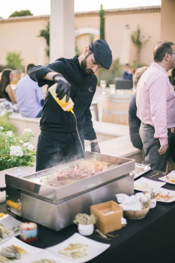 Uno chef che cucina carne alla griglia durante un evento all'aperto con persone sedute al tavolo.