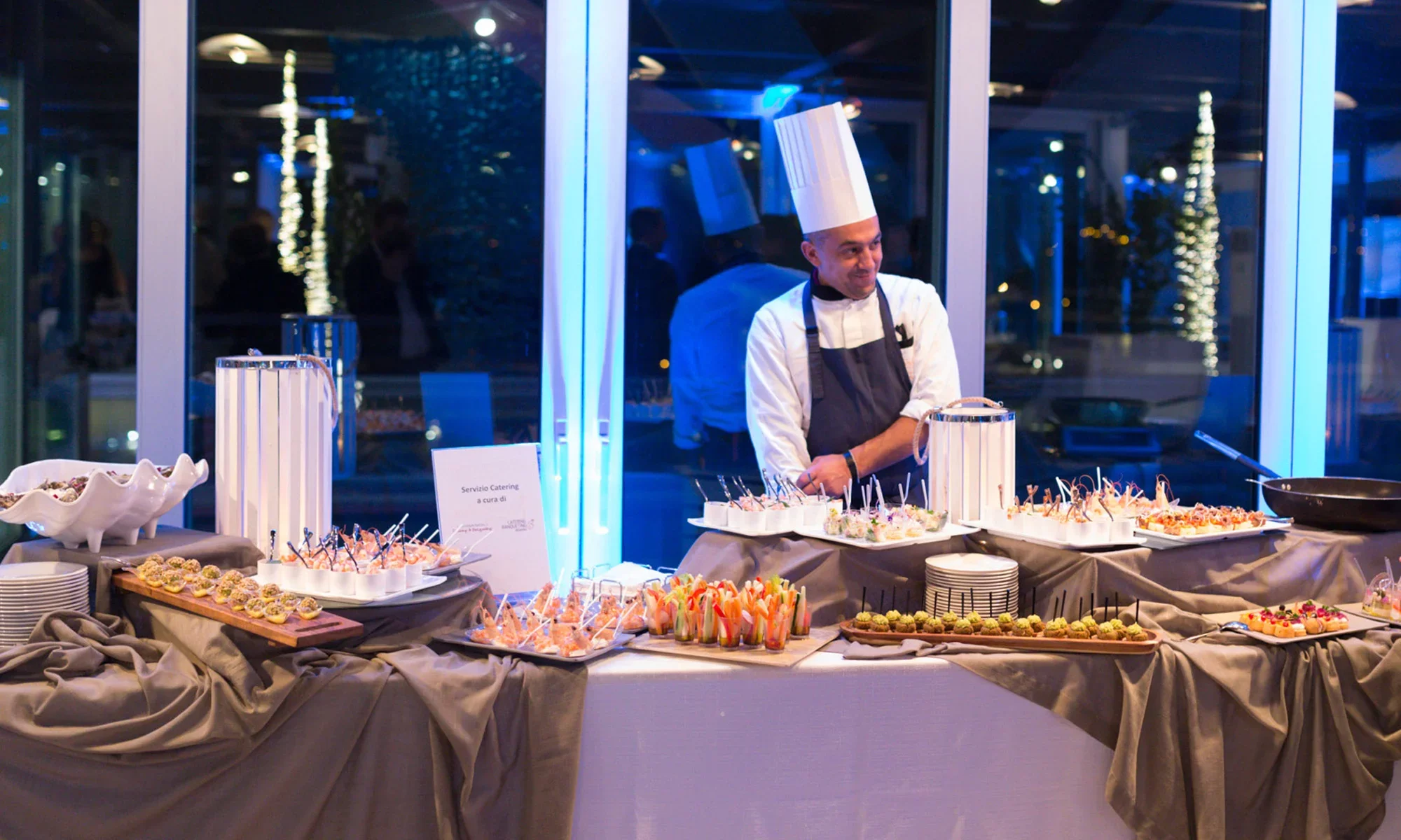 Una tavola con una vasta selezione di antipasti e finger food in un ambiente elegante, con un chef sorridente che supervisiona il servizio, atmosfera moderna e luci bluastre visibili attraverso le finestre.