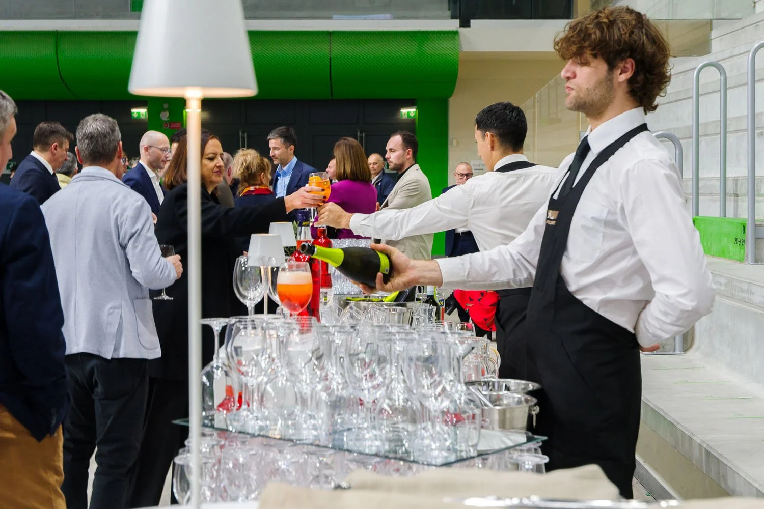 Gruppo di persone a un evento sociale che si scambiano drink e conversano, con un barista che versa vino in un bicchiere.