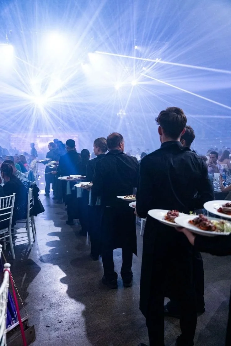 Persone a un evento sedute e in piedi, con alcuni camerieri che portano piatti di cibo, illuminato da luci e laser colorati