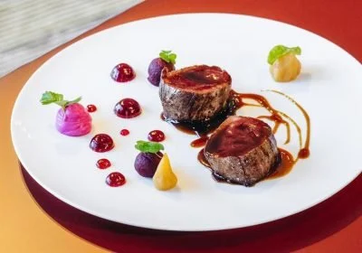 Due pezzi di carne con salsa, decorati con vari piccoli ciuffi di purè colorati e piccole sfere di frutta, presentati su un piatto bianco con bordo rosso.