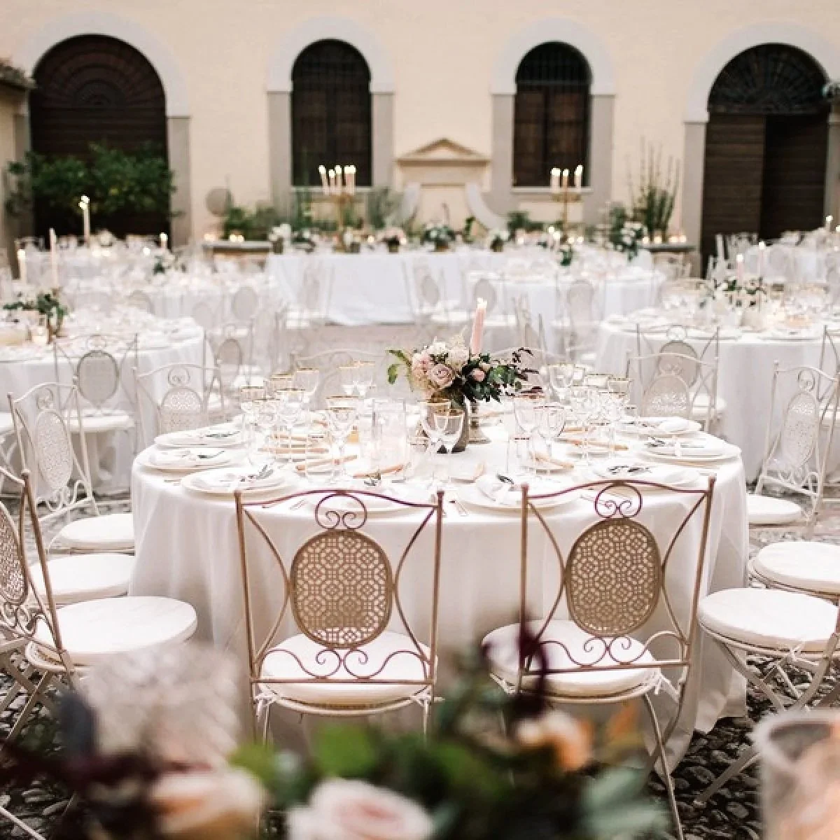 Tavola apparecchiata per un evento elegante con tovaglie bianche, centrotavola con fiori e candele, sedie di design bianco con dettagli dorati o color bronzo, ambientazione raffinata in un cortile o cortile interno.