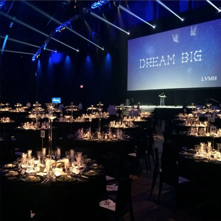 Sala da ballo elegante allestita per un evento con tavoli imbanditi e luci soffuse, sullo sfondo grande schermo con scritto "Dream Big" e il logo LVMH.