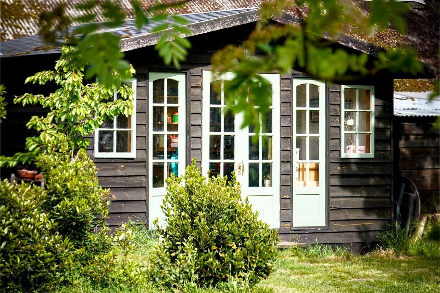 A small black wooden house with white framed French doors and windows, surrounded by green bushes and trees, with a grassy yard in front.