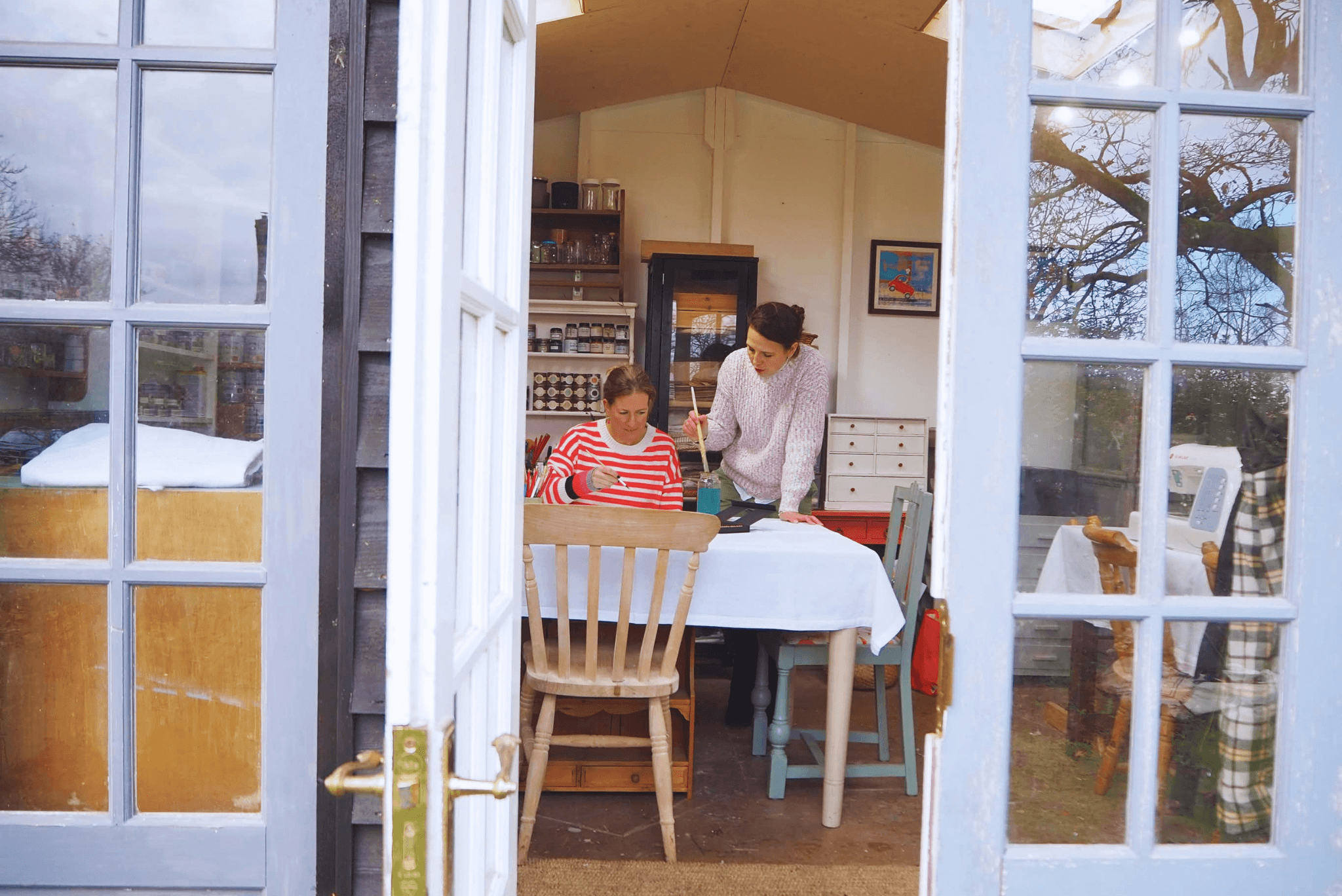 View through an open door showing a woman and a girl working at a table inside a cozy room with shelves of jars and glasses, with trees visible outside.