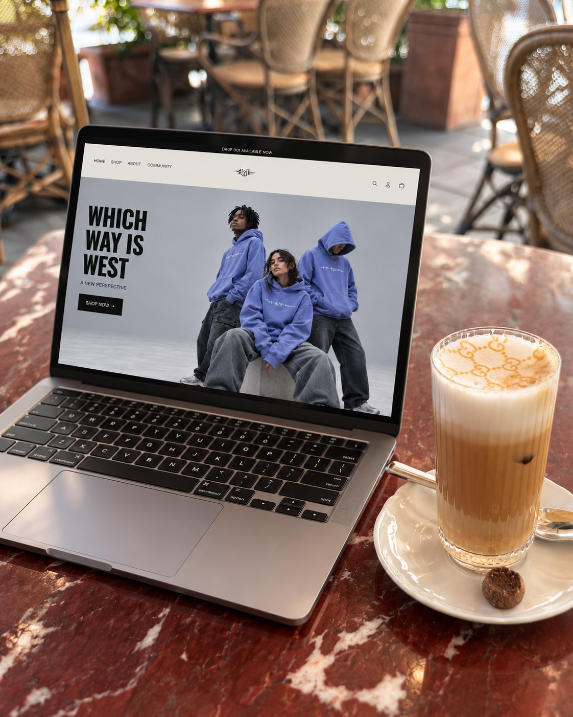 A laptop on a wooden table displaying a fashion website with three models wearing blue hoodies. Next to it is a glass of iced coffee with a straw, on a white saucer with a small chocolate cookie. In the background, there are wooden chairs and furniture in a bright outdoor setting.