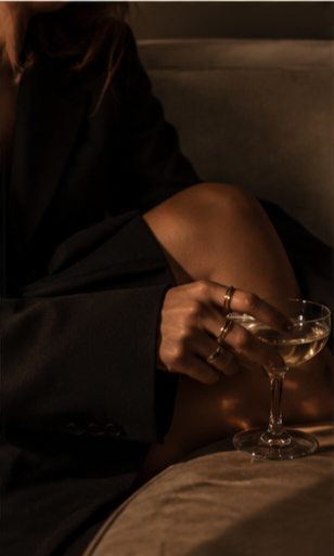 A person sitting on a bed holding a glass of champagne, wearing a black blazer with one leg showing.