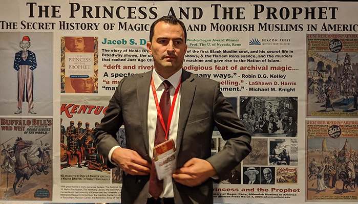 A man in a gray suit with a red tie and name badge stands in front of a newspaper-themed exhibition display. The display headliner reads 'The Princess and the Prophet' and discusses the secret history of magic and Muslims in America, featuring book covers, images, and text.