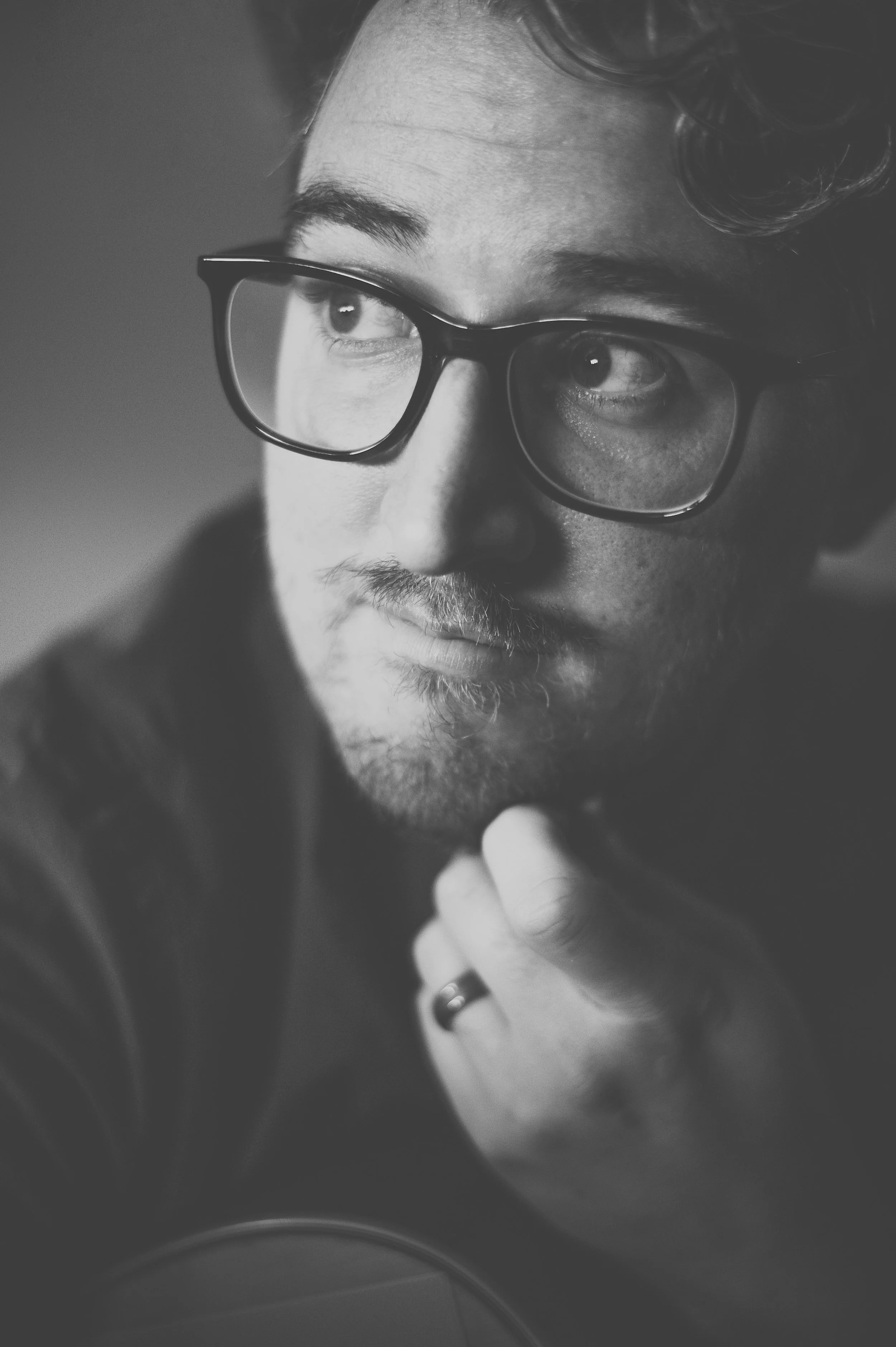 A black and white close-up photo of a man wearing glasses, looking pensively to the side, with a slight beard and mustache, holding his chin with his hand.