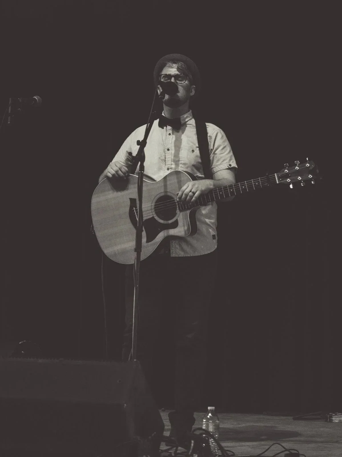 A musician performing on stage with an acoustic guitar, wearing glasses, a hat, a short-sleeved shirt, and a bow tie, in black and white.