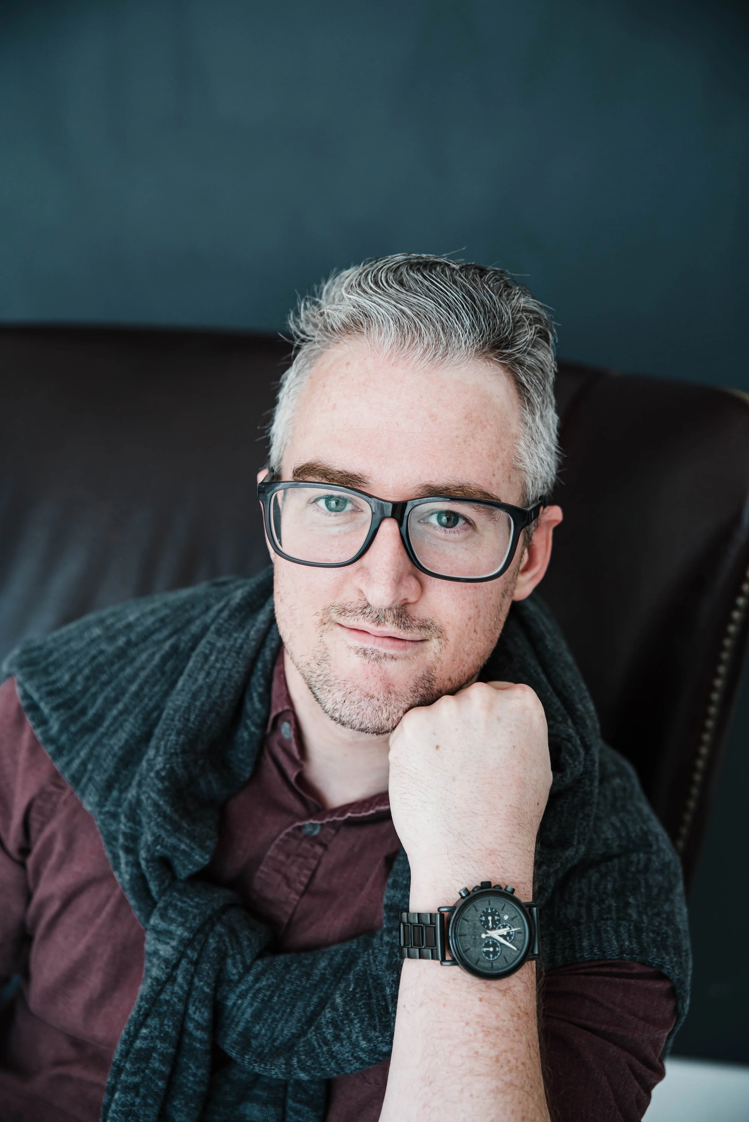 A man with gray hair and glasses sitting on a dark leather chair, looking at the camera with his chin resting on his hand. He is wearing a dark sweater over a maroon shirt and a black watch.
