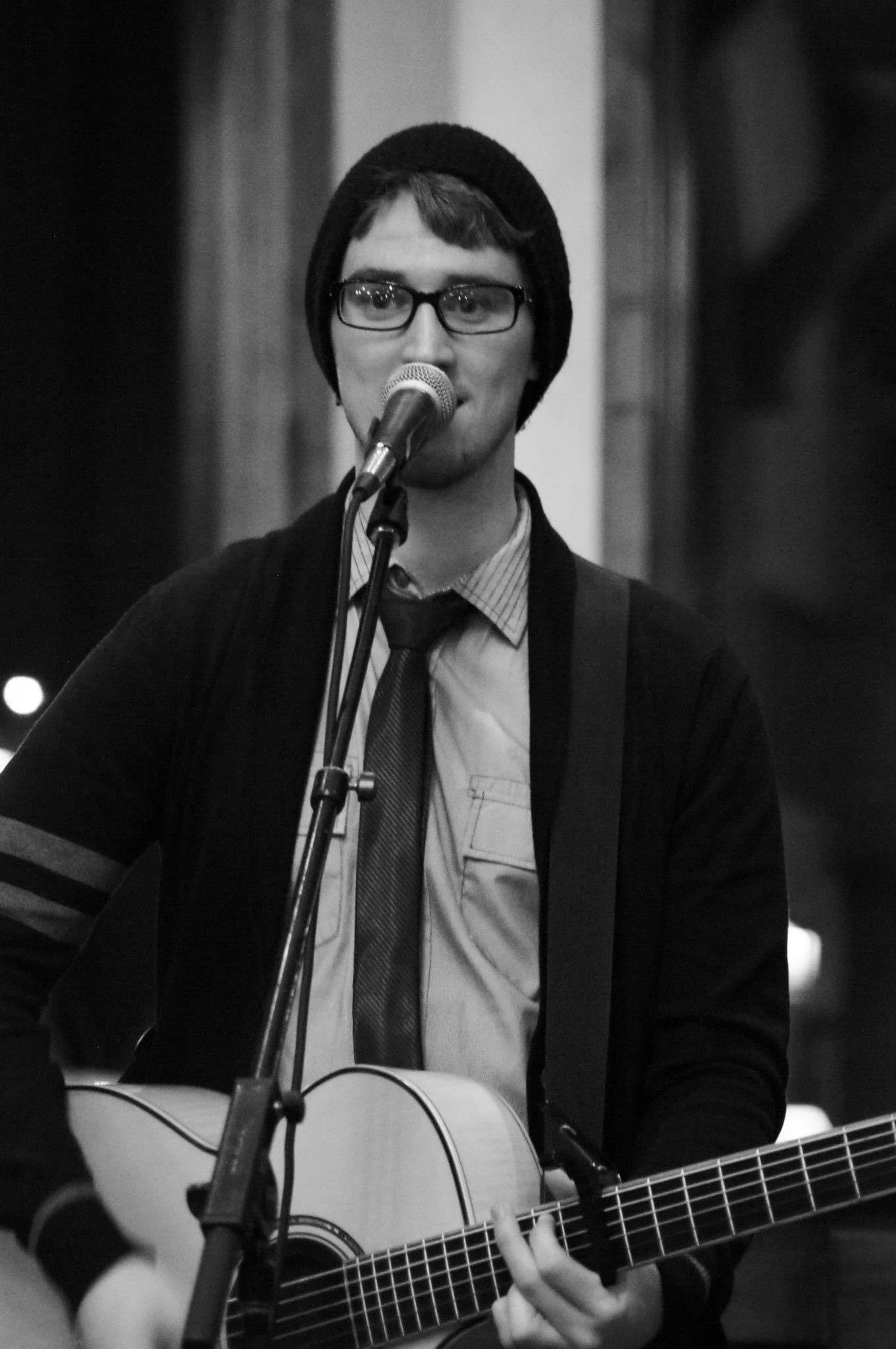 A young man with glasses and a beanie hat playing an acoustic guitar and singing into a microphone.